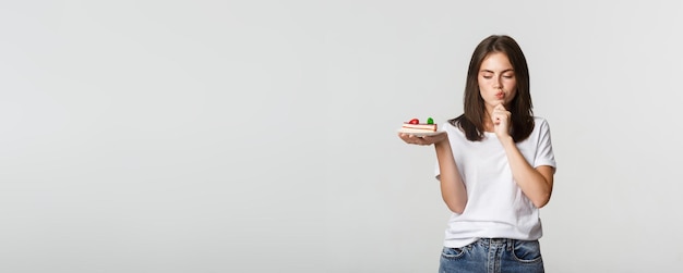 Foto gratuita chica guapa sonriente pensativa reflexionando mientras sostiene un trozo de pastel de fondo blanco.