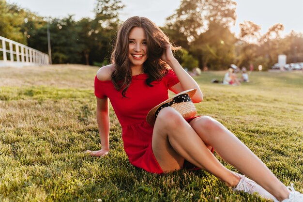 Chica guapa con sonrisa tímida sentada en el suelo en el parque. Mujer morena de buen humor en vestido rojo posando sobre la hierba en un día soleado.