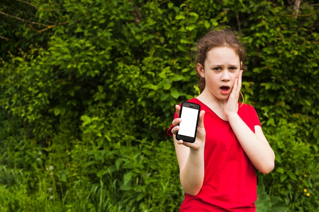 Chica guapa sorprendida mostrando teléfono celular de pantalla en blanco mientras está de pie en el parque