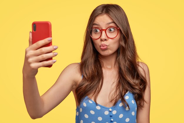 Chica guapa se toma selfie, hace pucheros en los labios del teléfono celular, hace videollamadas, coquetea con su novio, dispara algo para el blog, se toma una foto, usa lentes, modelos contra la pared amarilla