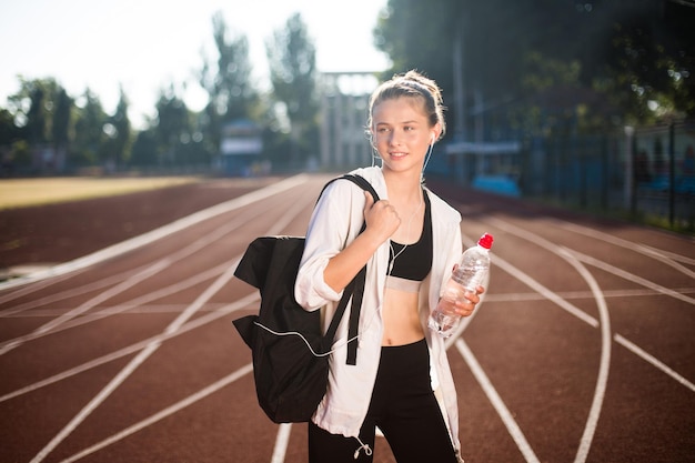 Chica guapa en top deportivo y legging mirando soñadoramente a un lado con mochila en el hombro y botella de agua pura en la mano en la pista de carreras del estadio