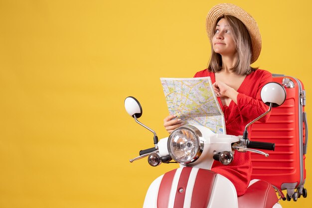 Chica guapa en vestido rojo en ciclomotor con maleta roja sosteniendo mapa