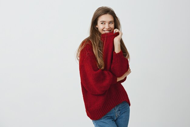 A la chica le gusta la ropa cómoda y suelta. Foto de estudio de mujer encantadora guapa en suéter rojo tirando del cuello mientras está parado a media vuelta, sonriendo y mirando a un lado con expresión coqueta