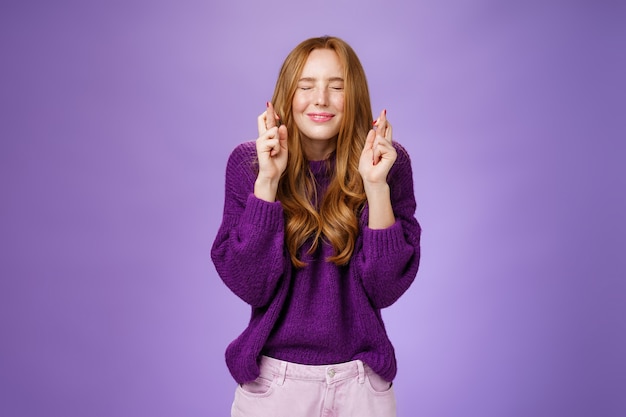 La chica hace que el deseo se haga realidad. Atractiva mujer joven pelirroja esperanzada y positiva en suéter púrpura con los ojos cerrados y sonriendo como dedos cruzados para la buena suerte, rezando y anticipando el milagro.