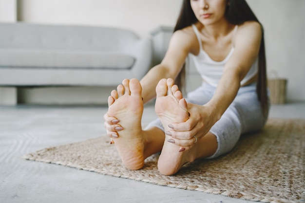 Chica haciendo ejercicios de yoga en casa cerca de un sofá y una ventana en ropa deportiva