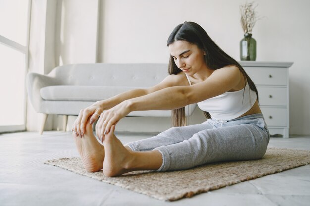 Chica haciendo ejercicios de yoga en casa cerca de un sofá y una ventana en ropa deportiva