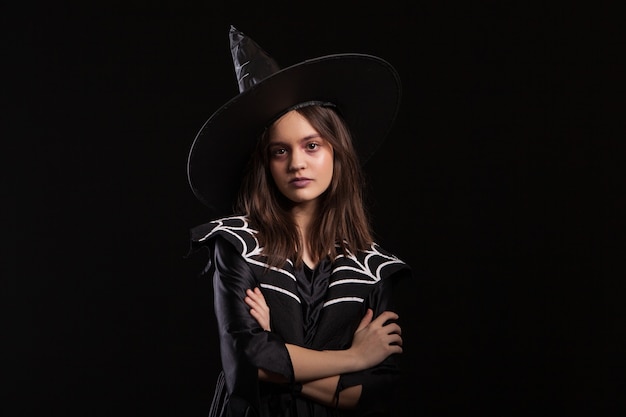 Chica haciendo hechizos oscuros con los brazos cruzados y expresión seria en el carnaval de halloween. Joven bruja haciendo brujería oscura.