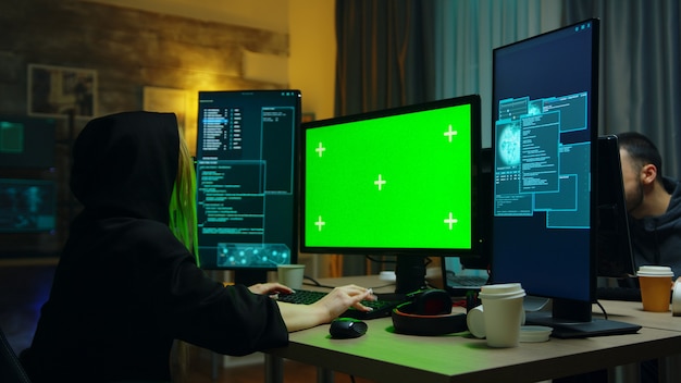 Chica hacker con una sudadera con capucha negra frente a la computadora con pantalla verde. Robo de identidad.