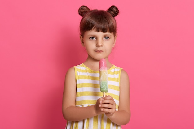 Chica con helado de agua posando aislada sobre fondo rosa, vestido de verano con rayas blancas y amarillas, de pie con divertidos nudos