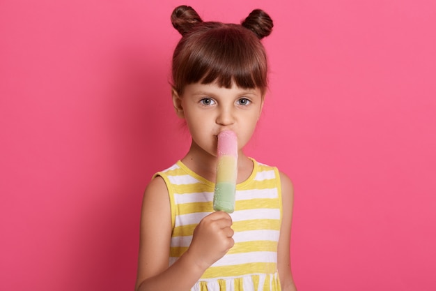 Foto gratuita chica con helado de agua, posando aislada sobre pared rosa, vistiendo atuendo blanco y amarillo a rayas, de pie con nudos divertidos.