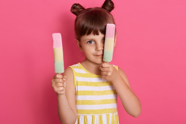 Chica con helados de agua en ambas manos posando aislada sobre pared rosa, cubriendo un ojo con sorbete, chica divertida con dos bollos de pelo y cuidado helado.