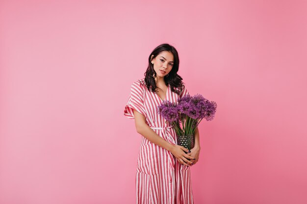 Chica hermosa atractiva en vestido de rayas posando con flores lilas. Morena se ve romántica.