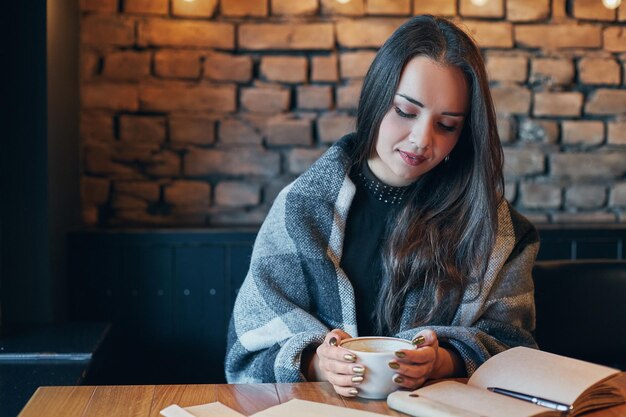 Una chica hermosa bebiendo café en un restaurante. Retrato de una joven con el cabello rizado oscuro cerrando los ojos soñadoramente con una taza en las manos. Una chica bonita sentada en un café con una taza de café