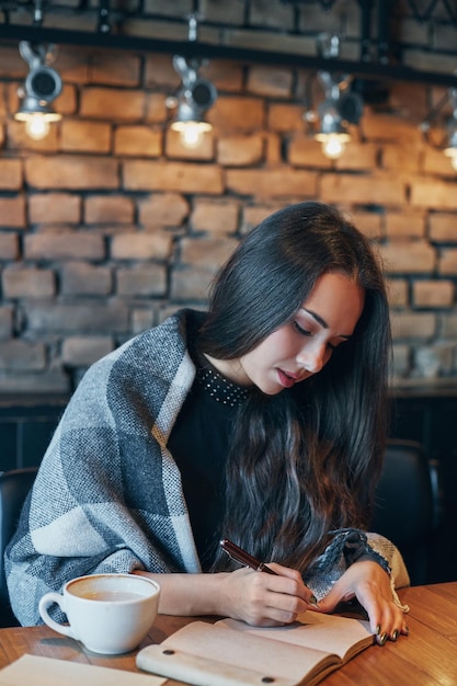 Foto gratuita una chica hermosa bebiendo café en un restaurante. retrato de una joven con el cabello rizado oscuro cerrando los ojos soñadoramente con una taza en las manos. una chica bonita sentada en un café con una taza de café