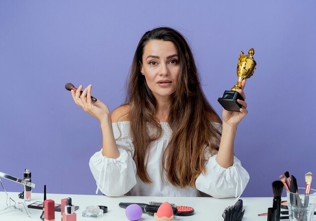 Chica hermosa confiada se sienta a la mesa con herramientas de maquillaje sostiene pincel de maquillaje y copa ganadora aislada en la pared púrpura