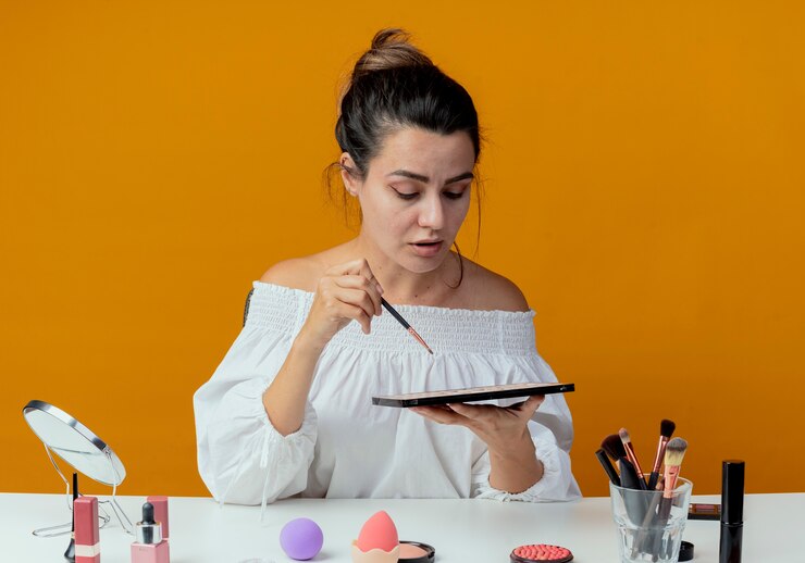 Foto gratuito chica hermosa confiada se sienta a la mesa con herramientas de maquillaje sostiene pincel de maquillaje y mira la paleta de sombra de ojos aislada en la pared naranja