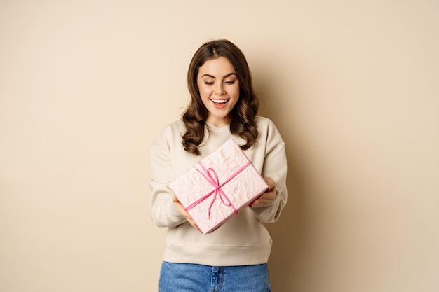 Una chica hermosa y emocionada con una caja de regalo envuelta en rosa, recibe regalos, de pie sobre un fondo beige.