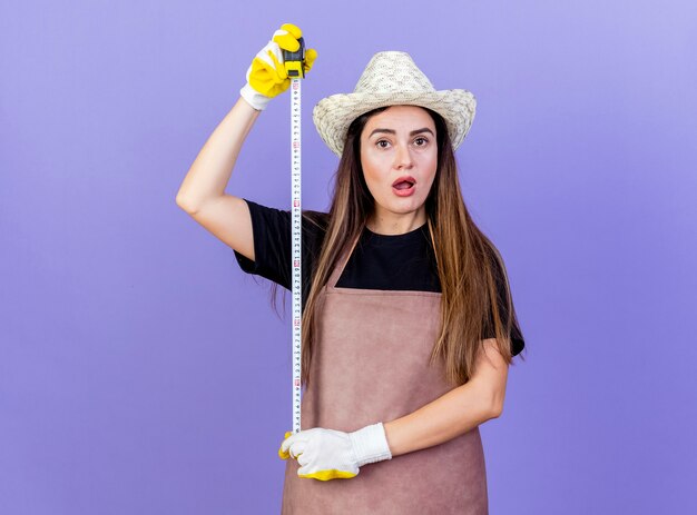 Chica hermosa jardinero sorprendida en uniforme con sombrero y guantes de jardinería que estira la cinta métrica aislada en azul