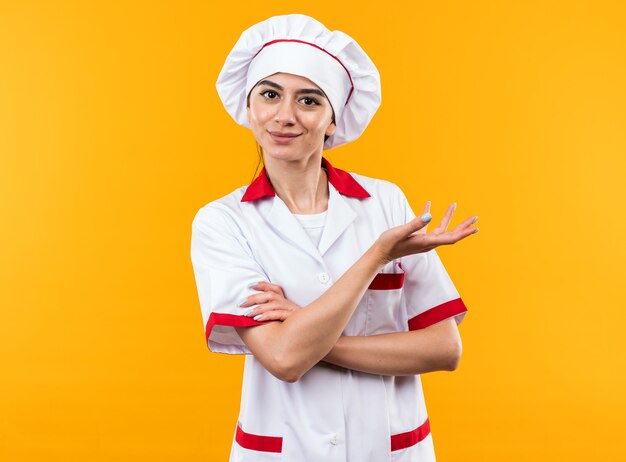 Chica hermosa joven complacida en puntos de uniforme de chef con la mano al lado aislado en la pared naranja