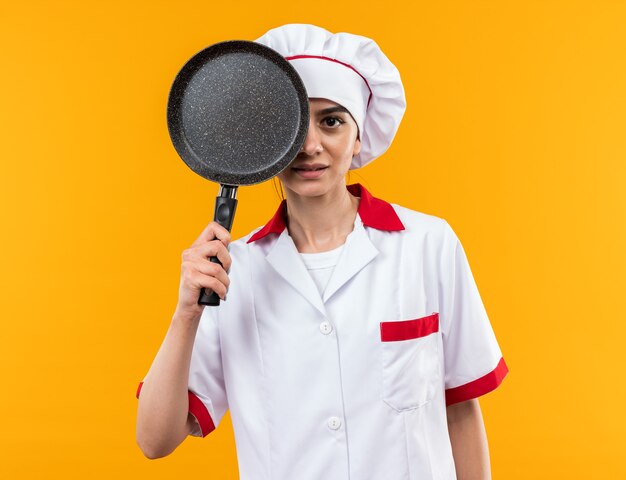 Chica hermosa joven complacida en uniforme de chef cubrió los ojos con una sartén