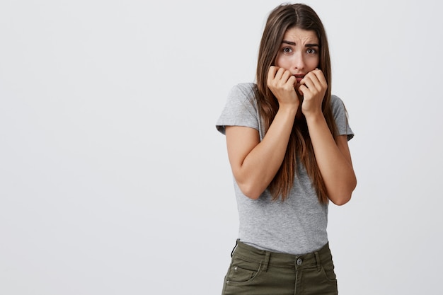 Chica hermosa joven infeliz estudiante caucásico con cabello largo castaño en camisa gris y jeans tomados de la mano cerca de la boca con expresión de miedo y preocupación.