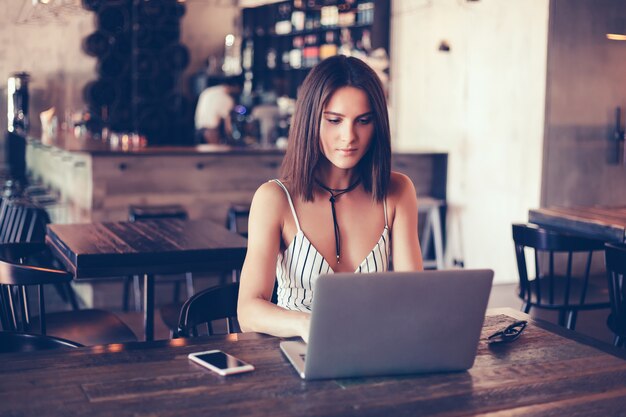 Chica hermosa joven usa una laptop en café, navegando en internet