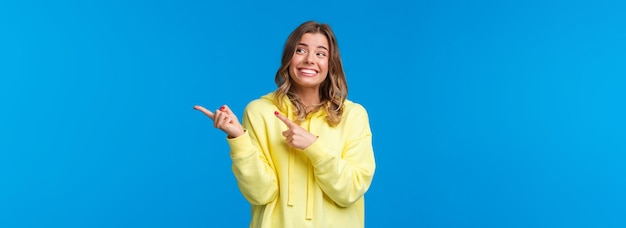 Chica hipster alegre con capucha amarilla con corte de pelo corto rubio apuntando y mirando hacia el lado derecho con