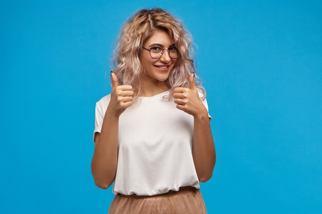 Chica hipster alegre en gafas redondas de moda haciendo gesto con el pulgar hacia arriba con ambas manos y sonriendo alegremente, mostrando su apoyo y respeto a alguien, diciendo Buen trabajo, bien hecho
