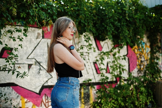 Chica hipster casual con estilo en gafas de gorra y jeans usa escuchar música de auriculares de teléfono móvil contra una gran pared de graffiti