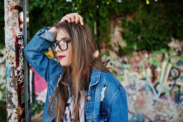 Chica hipster casual con estilo en jeans y gafas contra una gran pared de graffiti