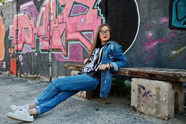 Chica hipster casual con estilo en jeans y gafas contra una gran pared de graffiti