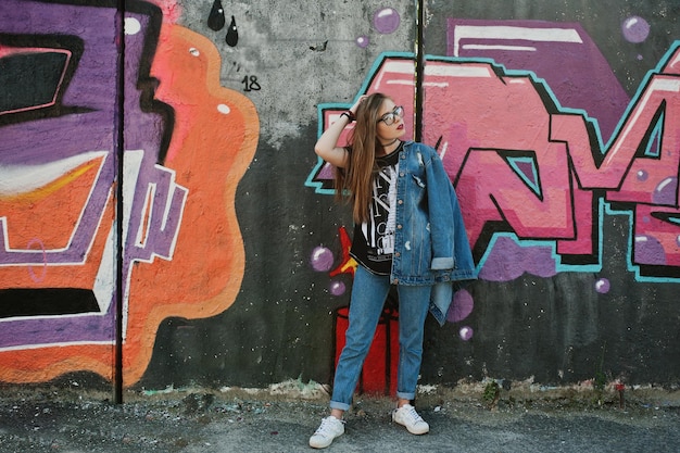 Chica hipster casual con estilo en jeans y gafas contra una gran pared de graffiti