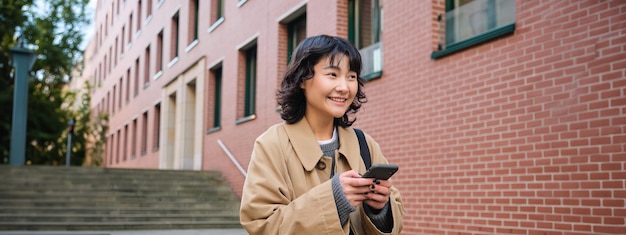 Una chica hipster coreana con estilo camina por el centro de la ciudad envía un mensaje de texto y utiliza una aplicación de mapa de ubicación en