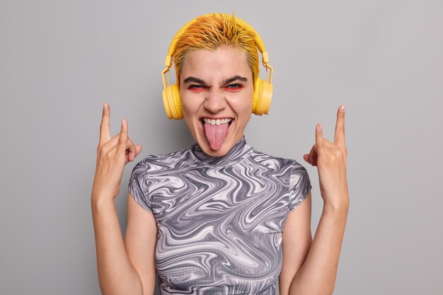 Chica hipster descarada con cabello amarillo de moda maquillaje vívido muestra cuernos de heavy metal gesto saca la lengua se divierte escucha música durante el tiempo libre de fiesta en el concierto disfruta del evento le gusta algo genial