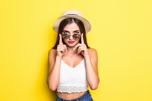 Chica hipster emocionada con sombrero y gafas de sol en la pared amarilla