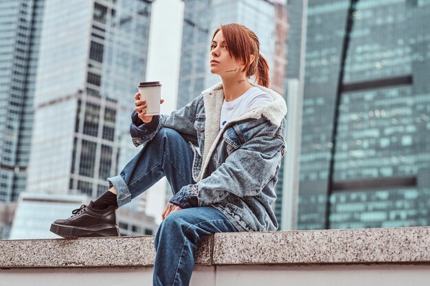 Chica hipster pelirroja elegante con tatuaje en la cara con abrigo de mezclilla sosteniendo café para llevar sentado frente a rascacielos en la ciudad de Moscú en la mañana nublada