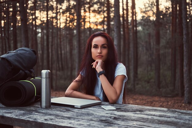 Chica hipster pensativa con camisa blanca mirando a la cámara mientras se sienta en un banco de madera con una laptop abierta en un hermoso bosque de otoño.