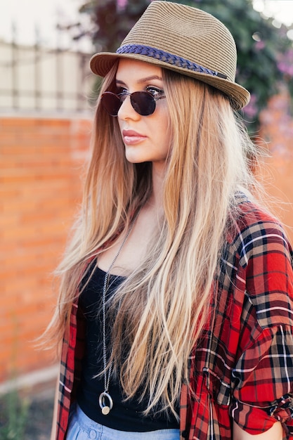 Chica hipster con sombrero retro y gafas de sol posando junto a la pared del grunge. Tiene el pelo largo, rubio y liso y camisa roja.