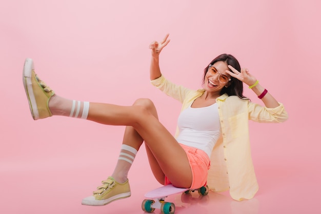 Chica hispana entusiasta en chaqueta amarilla de moda sentado en longboard con las piernas hacia arriba. Mujer latina alegre en coloridas pulseras riendo