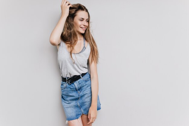 Chica impresionante bien formada jugando con el pelo largo y oscuro. Foto interior de adorable modelo de mujer caucásica en falda de mezclilla de moda.