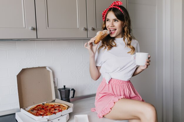 Chica increíble comiendo croissant juguetonamente en la mañana. Hermosa mujer rizada tomando café mientras está sentado al lado de la pizza.