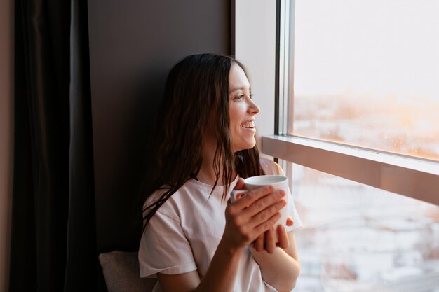 Una chica increíble, feliz y sonriente con el pelo oscuro y una camiseta blanca está sentada cerca de la ventana bajo el sol y bebiendo café por la mañana Chica con estilo en casa por la mañana