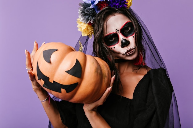 chica increíble con velo de novia negro aislado en la pared púrpura. Foto interior de la dama malvada en traje de zombi con calabaza de halloween.