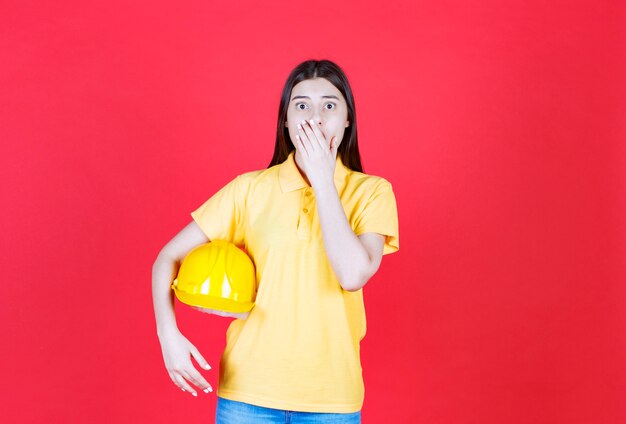 Chica ingeniera en código de vestimenta amarillo sosteniendo un casco de seguridad amarillo y parece aterrorizada y asustada.