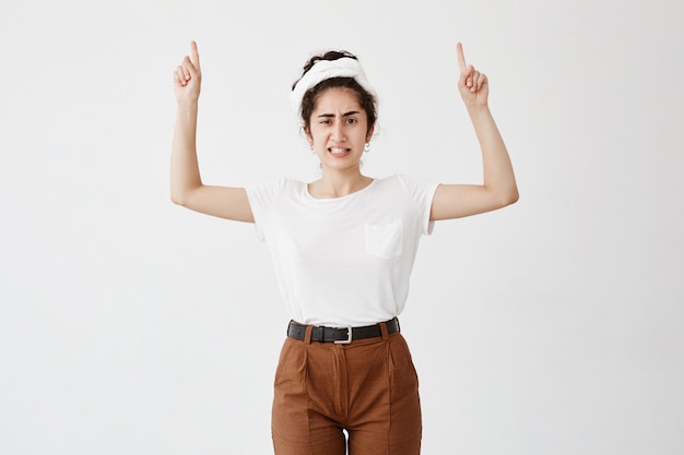 Chica insatisfecha con el pelo oscuro recogido en un moño, manteniendo los dedos apuntando hacia arriba, apretando los dientes blancos y frunciendo el ceño. Mujer joven vestida casualmente mostrando algo por encima de la cabeza, gestos con los dedos índices