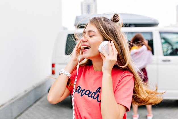 Chica inspirada con peinado de moda en elegante camisa rosa disfrutando de una buena canción con sonrisa y ojos cerrados