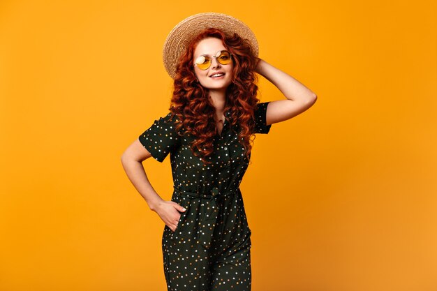 Chica inspirada en sombrero de paja posando con la mano en el bolsillo. Disparo de estudio de feliz sonriente mujer de jengibre en gafas de sol de pie sobre fondo amarillo.