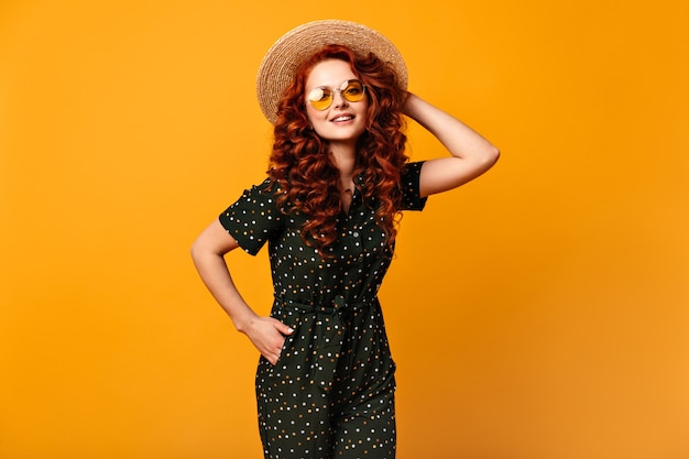 Chica inspirada en sombrero de paja posando con la mano en el bolsillo. Disparo de estudio de feliz sonriente mujer de jengibre en gafas de sol de pie sobre fondo amarillo.