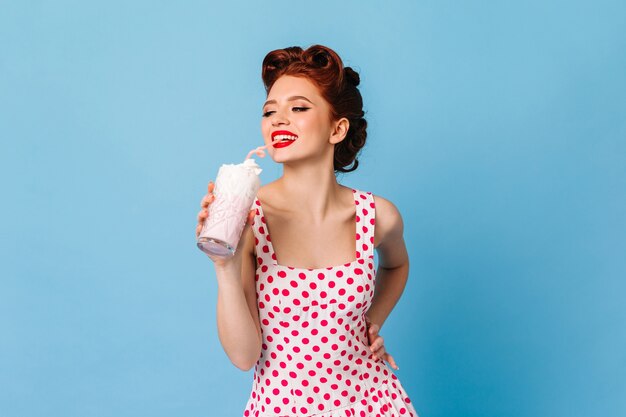 Chica inspirada en vestido de lunares bebiendo batido. Modelo femenino de jengibre riendo con bebida en el espacio azul.