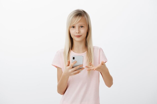 Chica inteligente respondiendo a una pregunta estúpida. Retrato de niña adorable descuidada con cabello rubio, gesticulando con la palma y sosteniendo el teléfono inteligente, sin darse cuenta y sin interés sobre la pared gris